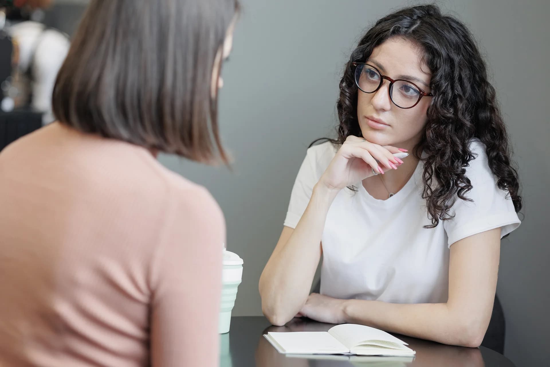 Mujer con gafas escucha a otra sentada frente a ella, con una libreta sobre la mesa
