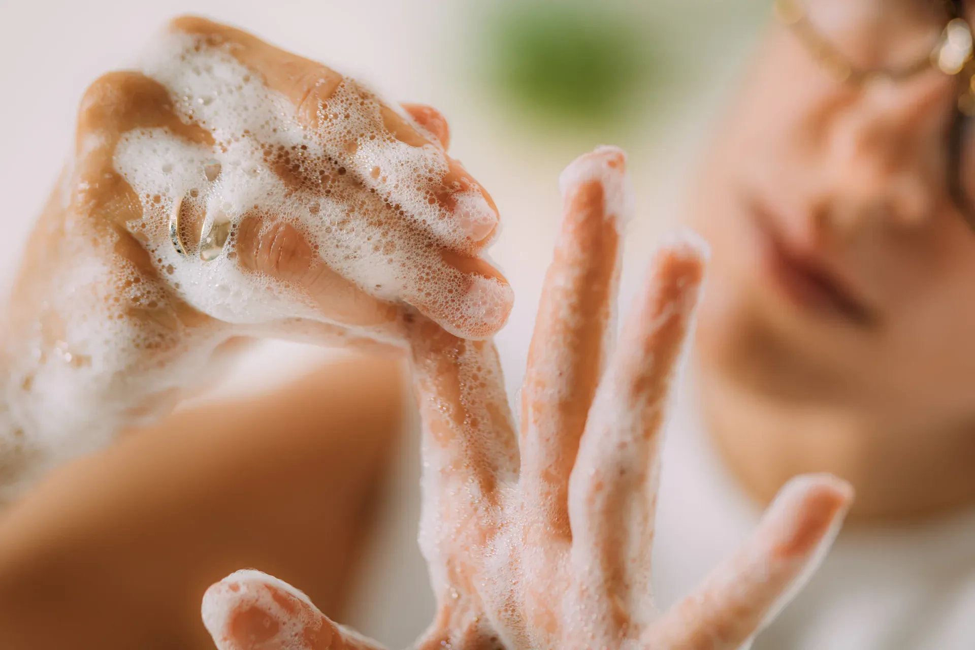 Manos cubiertas de espuma de jabón, con el rostro de una mujer desenfocado al fondo